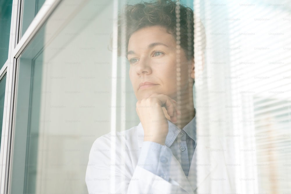 Young pensive female scientist or researcher in whitecoat keeping hand by chin while standing by window inside scientific laboratory