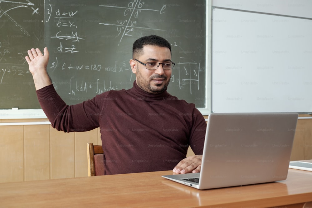 Mixed-race male teacher in casualwear pointing at solution of equation on blackboard while sitting by desk and looking at laptop display