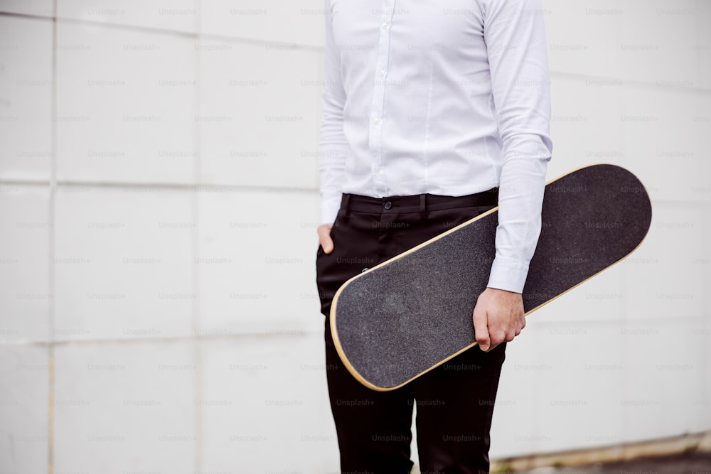 Closeup of man in shirt standing outdoors holding skateboard with hand in pocket.