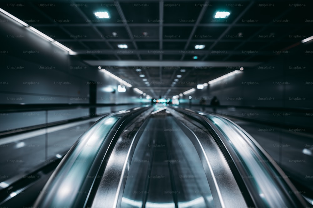 Blick mit geringer Tiefenschärfe von zwei Fahrgästen in einen modernen Flughafen mit selektivem Fokus im Vordergrund; Zwei Laufbänder in einem dunkelblauen modernen Terminalinnenraum in der Nähe von Flugsteigen