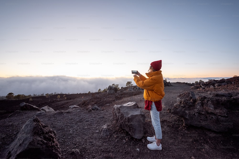 Woman in bright jacket and hat phototgraphing on phone beautiful sunset cloudscape, traveling higly on the rocky mountains