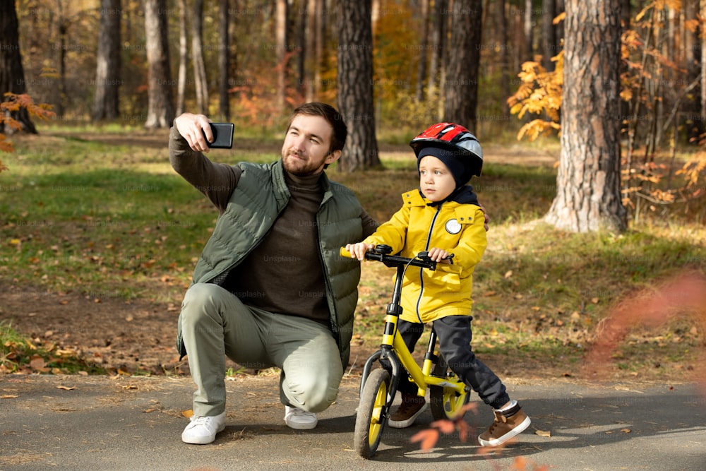Happy young bearded man with smartphone making selfie with his cute little son in safety helmet sitting on balance bicycle in public park