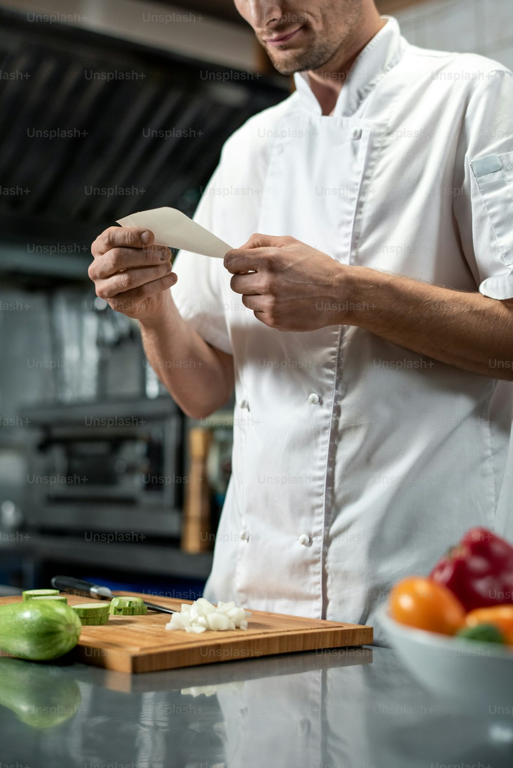 Young chef in uniform looking through list of ingredients for vegetable salad while holding paper over chopped onion on wooden board