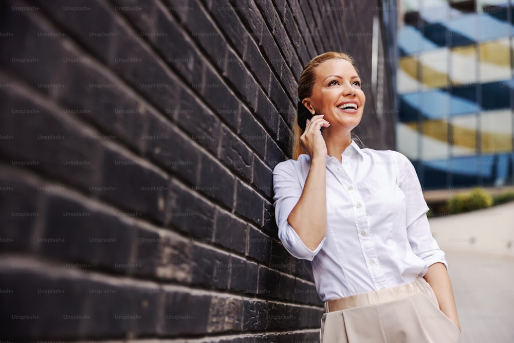 Smiling cheerful fashionable blond businesswoman leaning on the wall outdoors and talking on the phone.
