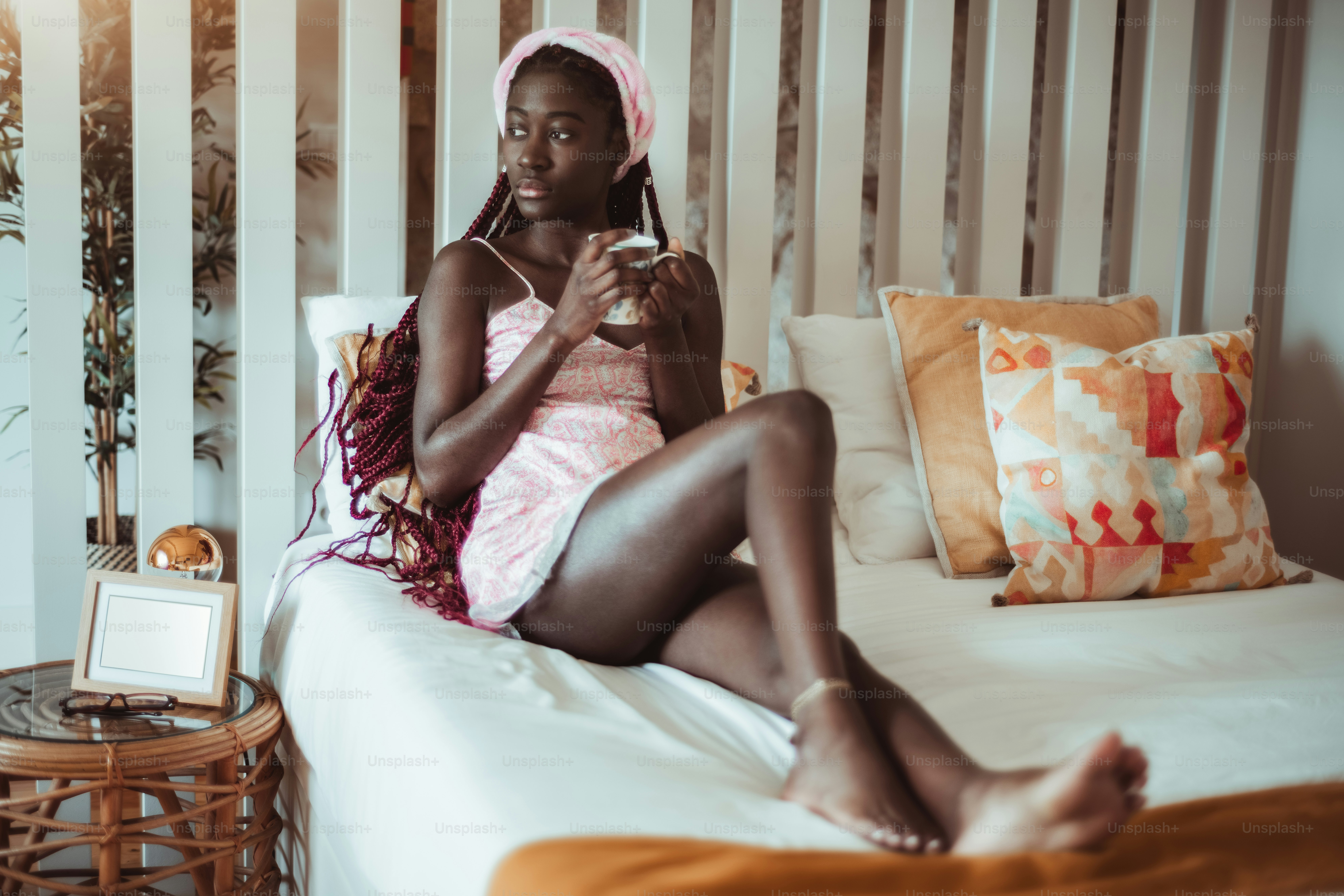 ベッドガウンを着て三つ編みをした裸足の若い女性が、居心地の良いアパートの寝室のベッドで、おいしいお茶を手に持って脇を見ながら、物思いにふけっている