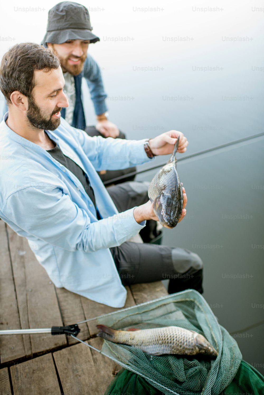 Two happy fishermen holding caught fish sitting on the wooden pier during the fishing on the lake at the morning