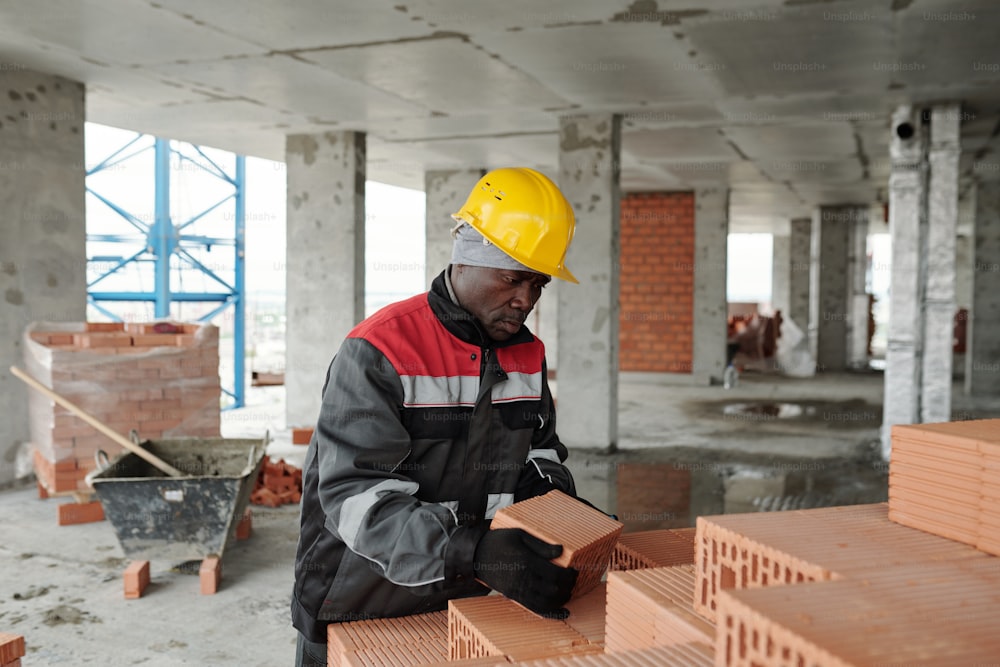 작업복과 보호 헬멧을 쓴 성숙한 흑인 남성이 건설 현장의 미완성 건물에서 일하는 동안 벽돌을 쌓아 올립니다.
