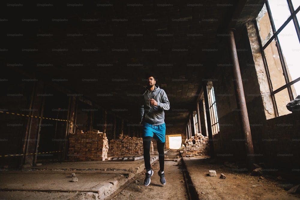 Portrait of active motivated afro-american young attractive athletic man with earphones running inside of the abandoned place.