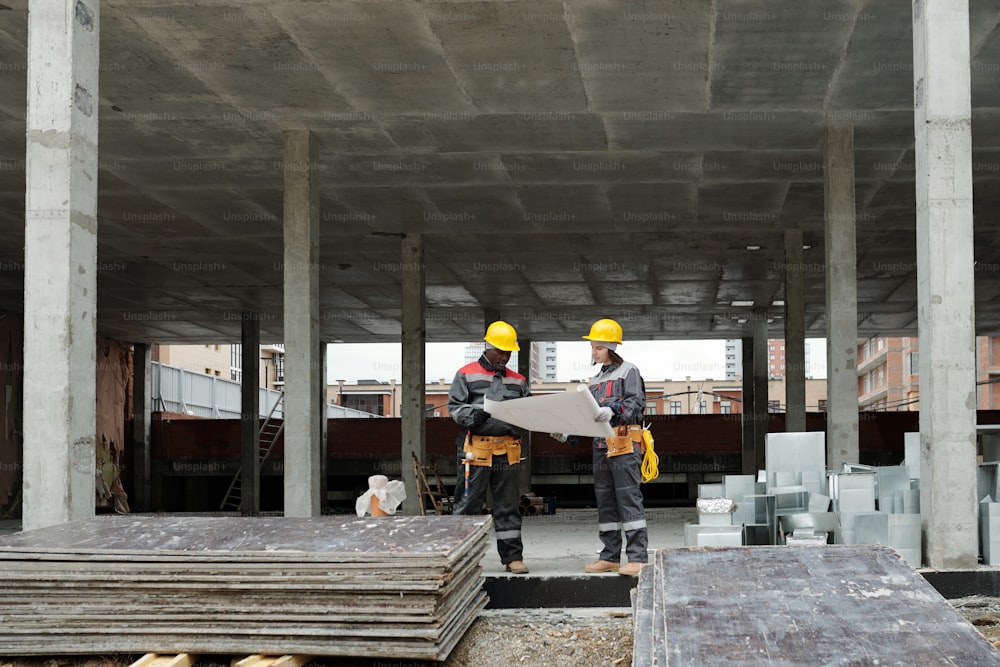 Dois construtores interculturais modernos em roupas de trabalho discutindo o plano arquitetônico enquanto seguram a planta desenrolada na área de construção