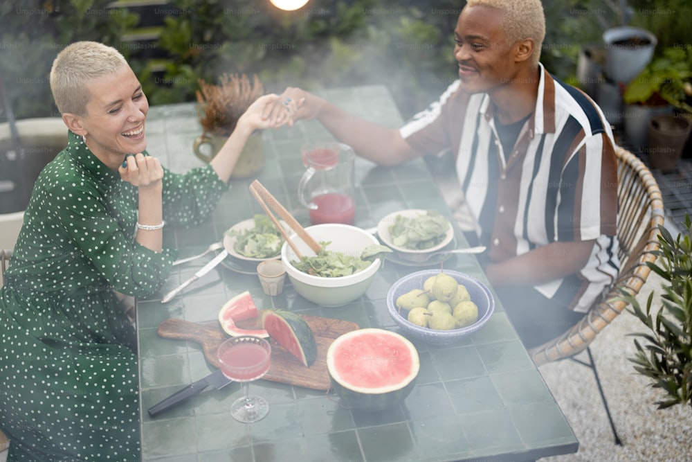 Multirassisches Paar beim Abendessen im Hinterhof ihres Landhauses in der Natur. Idee von gesunder Ernährung und modernem Lebensstil. Schwarzer Mann und Europäerin genießen die gemeinsame Zeit