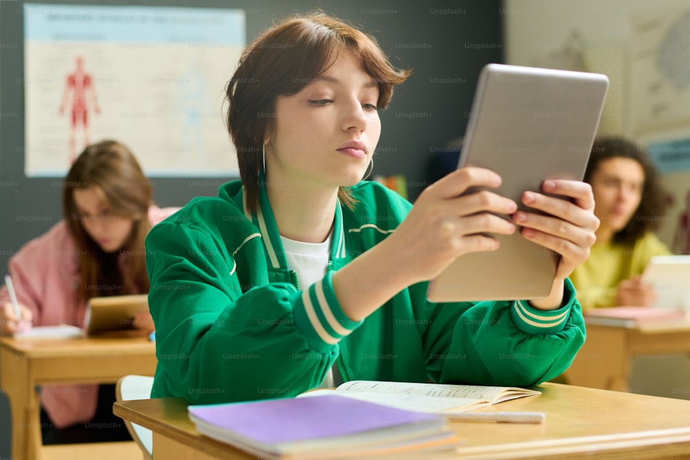 Serious female student of highschool holding tablet in front of her face while sitting by desk and looking through online information at lesson
