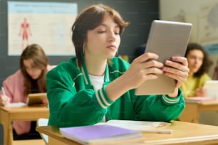 Serious female student of highschool holding tablet in front of her face while sitting by desk and looking through online information at lesson