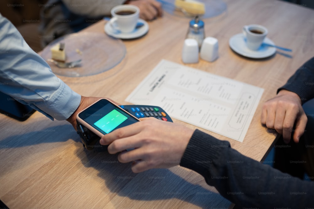 Joven sosteniendo el teléfono inteligente sobre el terminal de pago sostenido por la mano de la camarera mientras está sentado junto a la mesa de madera en el café