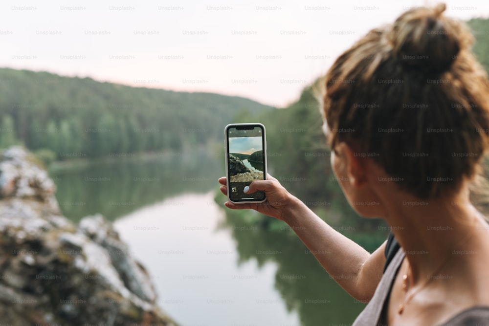 Mujer joven delgada con mochila tomando foto de la hermosa vista de las montañas y el río tranquilo en el teléfono móvil, viaje local
