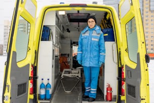 Young female paramedic in uniform standing by empty stretcher inside ambulance car in front of camera