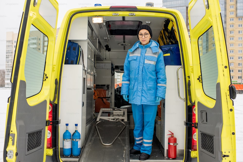 Young female paramedic in uniform standing by empty stretcher inside ambulance car in front of camera