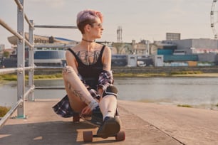 Portrait of young pink haired tattooed girl in round glasses sitting on a longboard at the river bank with city view, looking away from the camera