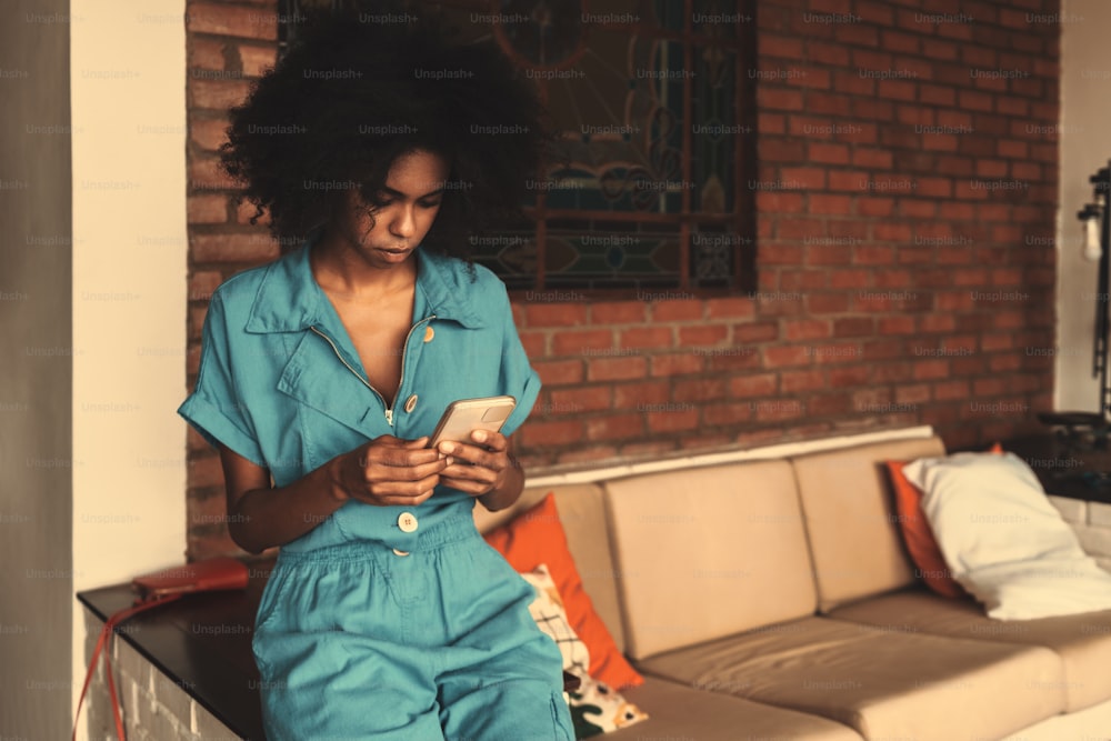 A portrait of a dazzling Brazilian female in a teal overalls sitting on the side of the sofa indoors at home and reading incoming e-mail on the screen of her phone; a copy space place on the right