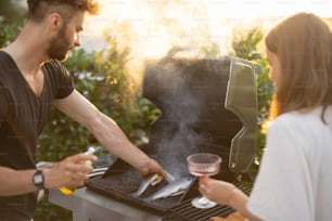 Hombres cocinando trucha fresca en una moderna parrilla de gas al aire libre al atardecer. Cocinar alimentos saludables al aire libre