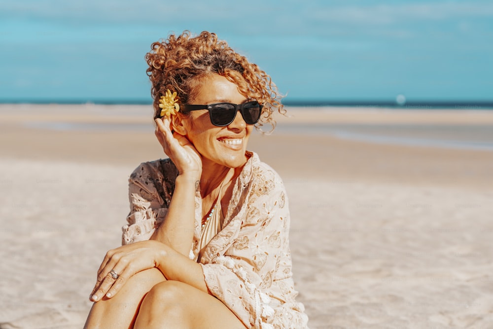 Portrait of happy tourist sitting and smiling at the beach with blue sky and ocean in background. Travel and tourism in summer holiday vacation. Female people with sunglasses on the sand