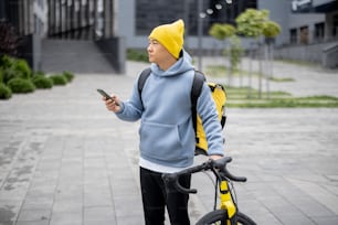 Kurier mit Fahrradsuche Kundenplatz mit GPS auf dem Smartphone in der Stadt. Konzept des Versands und der Logistik während der Coronavirus-Pandemie. Vorstellung von Beruf und Job. Asiatischer Mann in warmer Kleidung