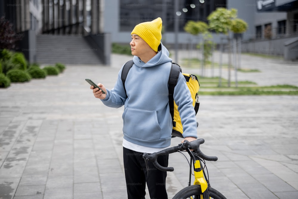 Courier with bicycle searching client's place with GPS on smartphone in city. Concept of shipping and logistics during Coronavirus pandemic. Idea of profession and job. Asian man wearing warm clothes