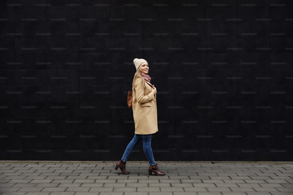 여성을위한 겨울 스타일과 패션. 코트, 스카프, 비니를 입은 젊은 웃는 여자의 전체 길이가 거리를 걷고 회색 벽을 지나갑니다.