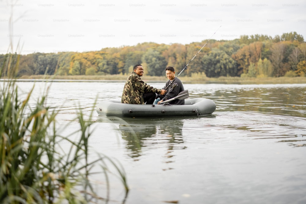 Multiracial male friends fishing with fishing rods on rubber boat in lake or river. Concept of rest, weekend and hobby in nature. Idea of friendship and spending time together. Autumn day