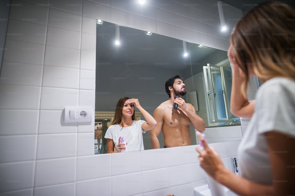 Beau jeune et beau couple d’amour souriant debout ensemble devant le miroir et préparant leur peau pendant que l’homme tient une tondeuse à cheveux le matin à la salle de bain.