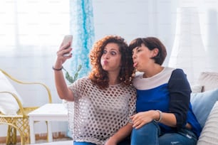 自宅で携帯電話で写真を撮る2人の若い女性が、素敵で面白い表情をしています。友達とネット上でコミュニケーションをとるのが大好きな親友。