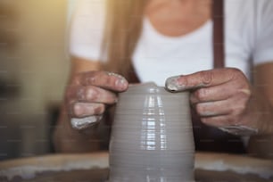 Creative artisan shaping wet clay with her hands on a pottery wheel while sitting in her ceramic workshop