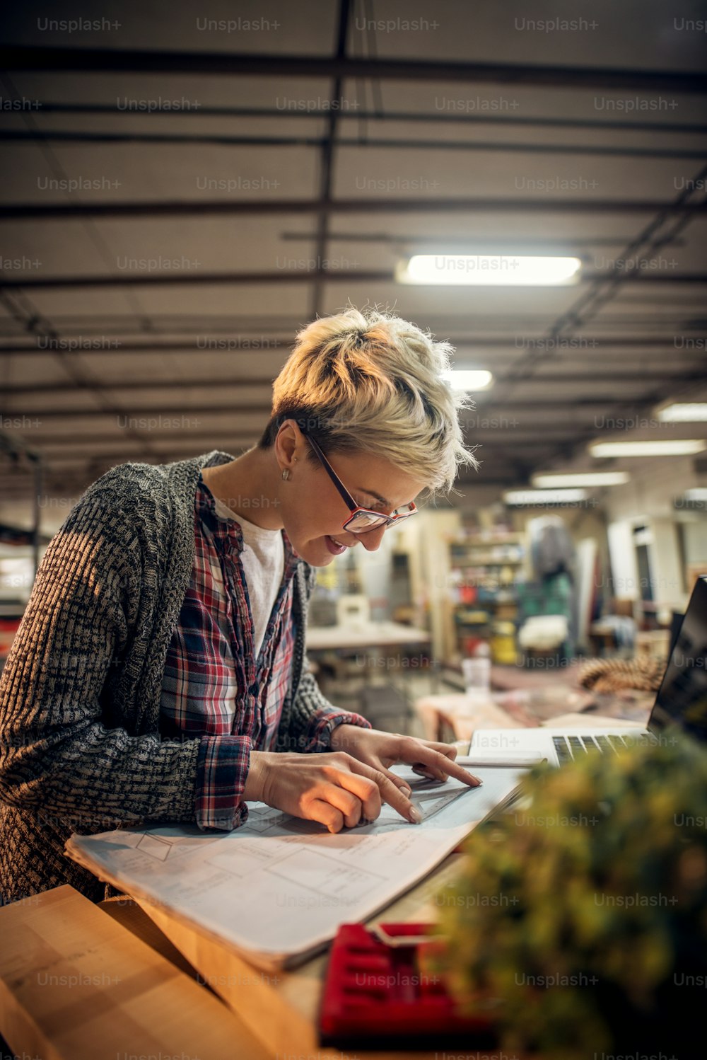 Close up view of charming smiling motivated short hair attractive middle aged industrial female engineer with eyeglasses working with blueprints and laptop in the workshop.