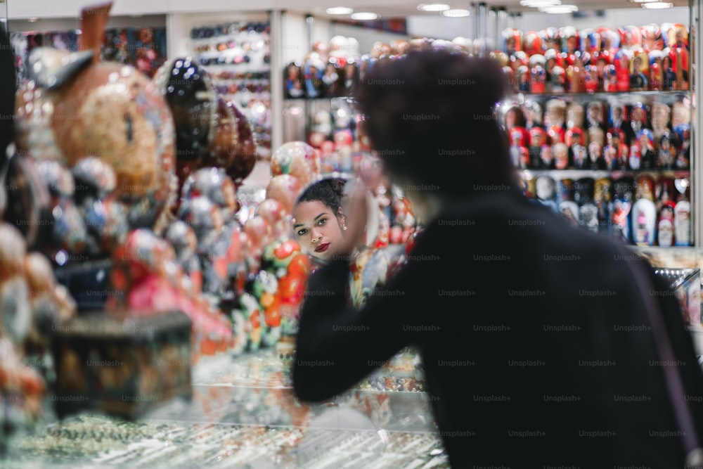 Giovane turista afroamericana sta guardando in un piccolo specchio rotondo all'interno di un negozio di souvenir e fissando i suoi capelli circondati da un sacco di souvenir colorati sfocati, messa a fuoco selettiva sul suo viso