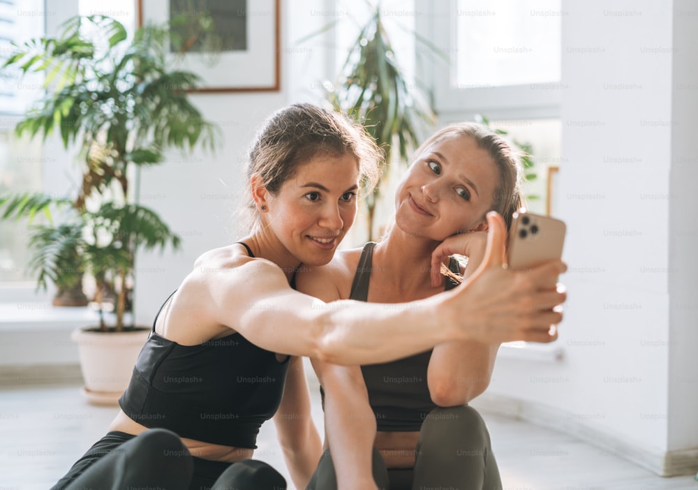 Young fit women friends practice yoga taking selfie on mobile phone in a yoga studio