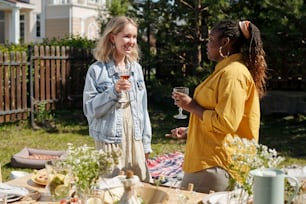 Young black woman and blond Caucasian girl with glasses of homemade wine having chat during outdoor party by served table