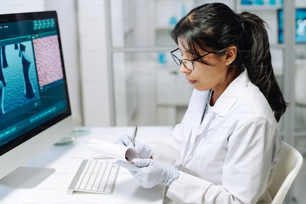 Young serious Hispanic female scientist making notes in her notepad while sitting by workplace in front of computer during research