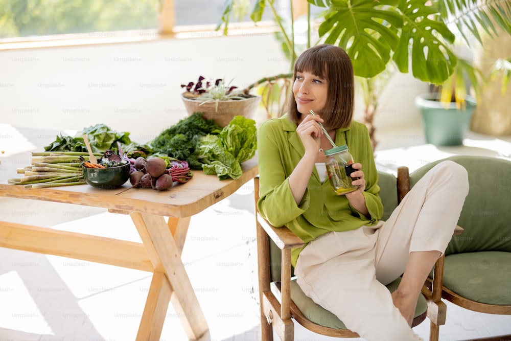 Una mujer joven bebe limonada mientras está sentada en una silla cerca de la mesa con muchos ingredientes de alimentos frescos en la habitación con plantas verdes. Concepto de estilo de vida saludable