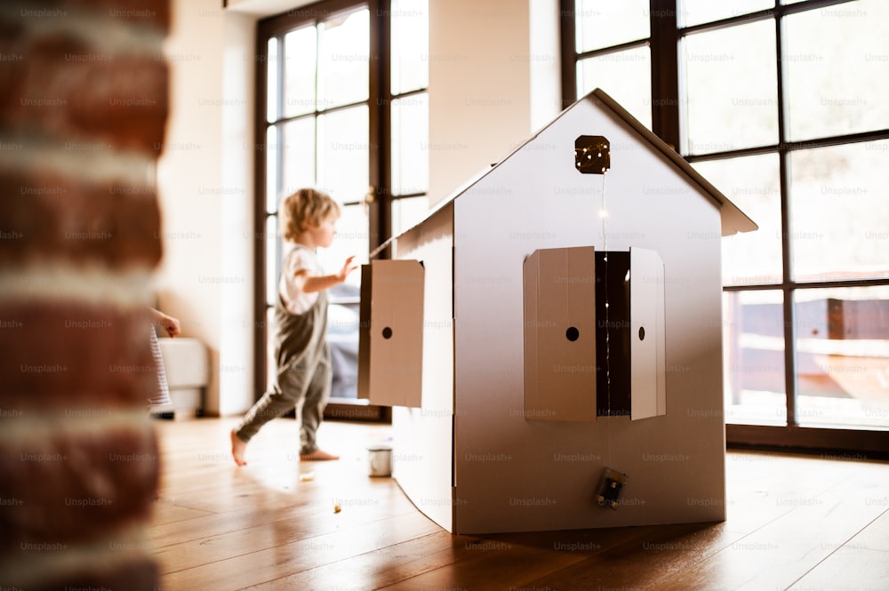 A happy toddler boy playing with a carton paper house indoors at home.