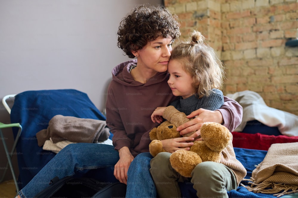 Young woman giving hug to her cute little son with brown soft teddybear while both sitting on sleeping place prepared for refugees