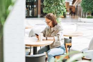 Giovane studentessa adolescente bruna in occhiali usando il telefono cellulare al caffè moderno verde, spazio aperto del luogo pubblico