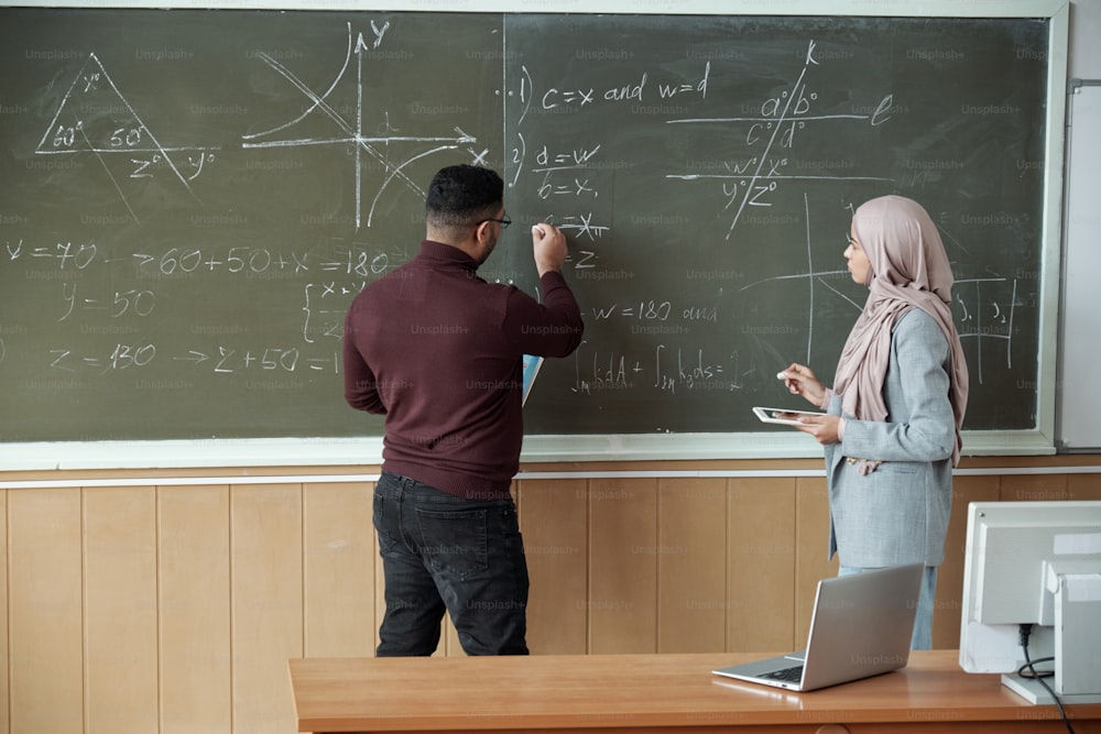 Male professor and young female student in hijab standing by blackboard and solving equation at lesson while man pointing at one of derivatives