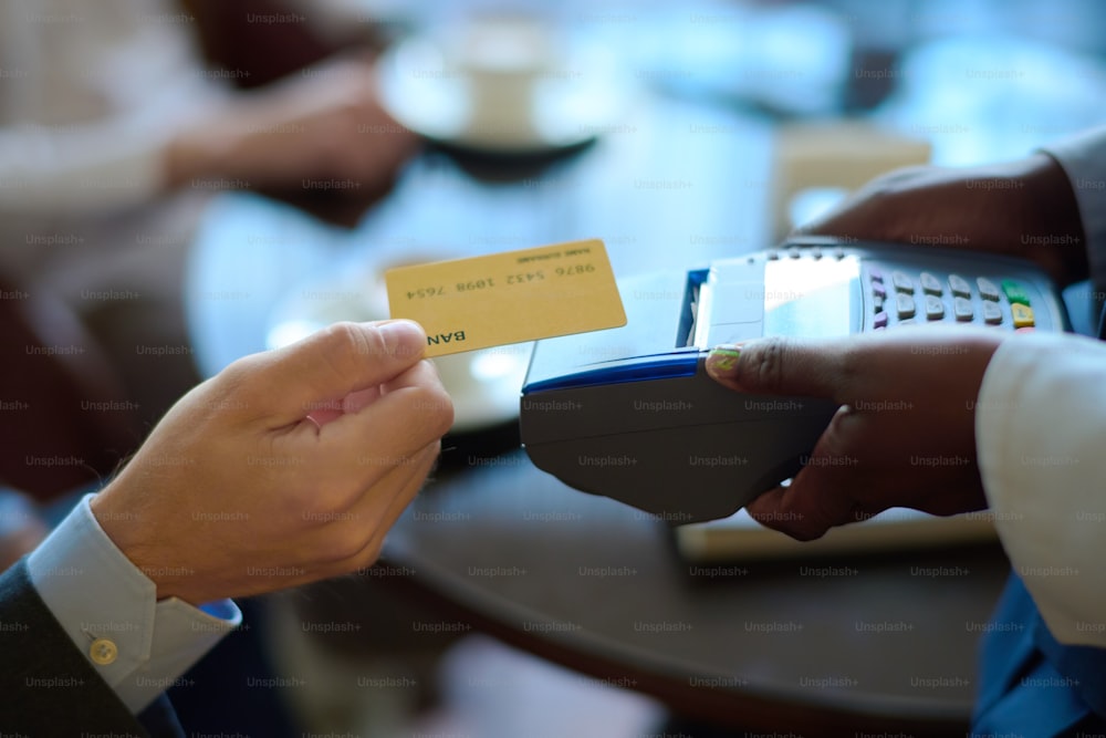 Hand eines jungen Geschäftsmannes, der die Kreditkarte über dem Zahlungsterminal hält, das von einer afroamerikanischen Managerin des modernen Hotels gehalten wird