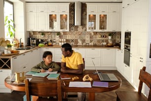 Young father and his son communicating during homework while sitting by wooden table in the kitchen