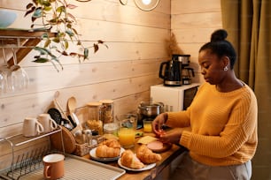 Contemporary young blaxck woman in yellow knitted sweater preparing sandwiches with cheese, ham and fresh chopped tomatoes
