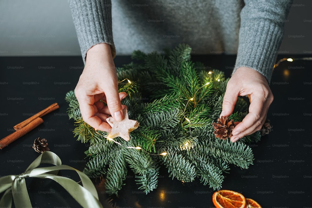 Junge Frau in warmem grauem Strickkleid macht DIY-Tannen-Weihnachtskranz in der Hand auf dem Tisch zu Hause