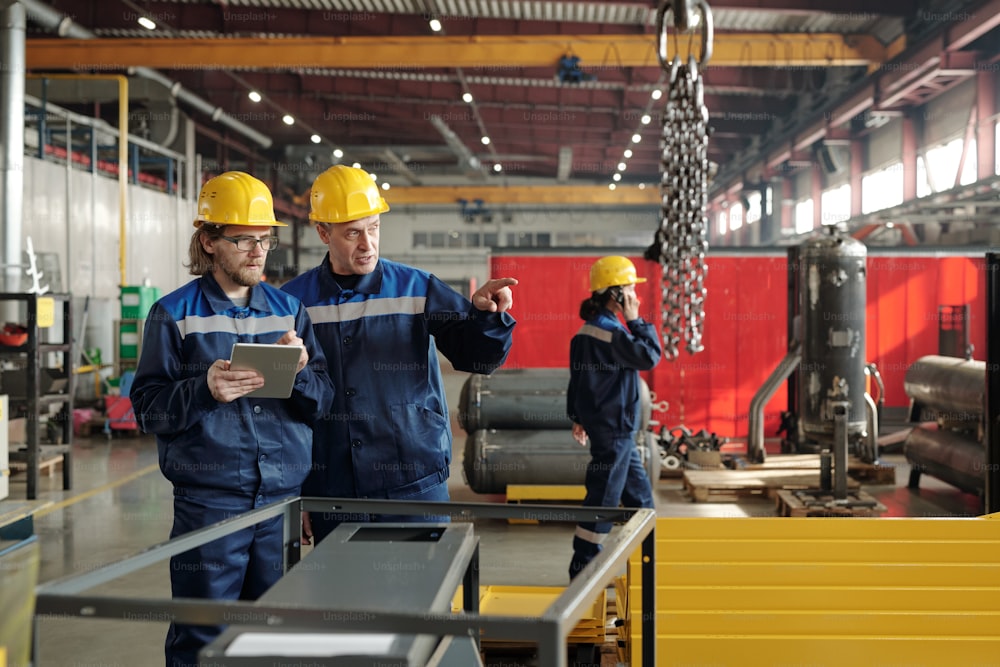Two engineers in workwear standing in large workshop or plant and discussing new industrial equipment while one of them pointing at it
