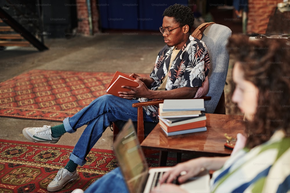 Serious African American guy in stylish shirt and blue jeans opening book in brown cover while sitting in armchair in living room at leisure