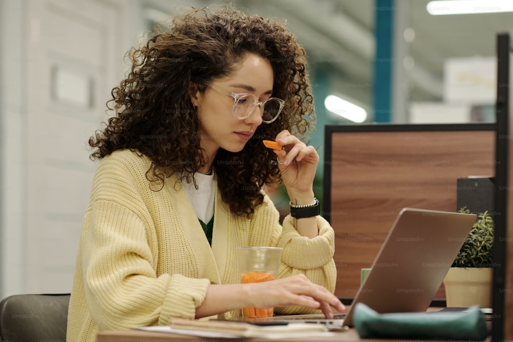 Young serious woman eating fresh carrots while sitting by workplace in front of laptop and concentrating on network in openspace office
