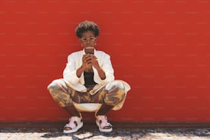 Uma jovem africana deslumbrante e extravagante está agachada em frente a uma parede vermelha e usando seu smartphone; uma mulher negra elegante de óculos escuros, calças de camuflagem e nail art está telefonando perto de uma superfície vermelha