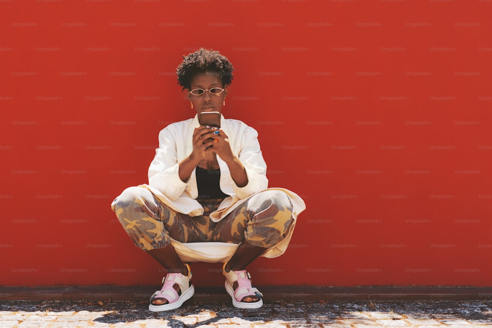 A dazzling fancy young African female is squatting in front of a red wall and using her smartphone; an elegant black woman in sunglasses, camouflage pants, and nail art is phoning near a red surface